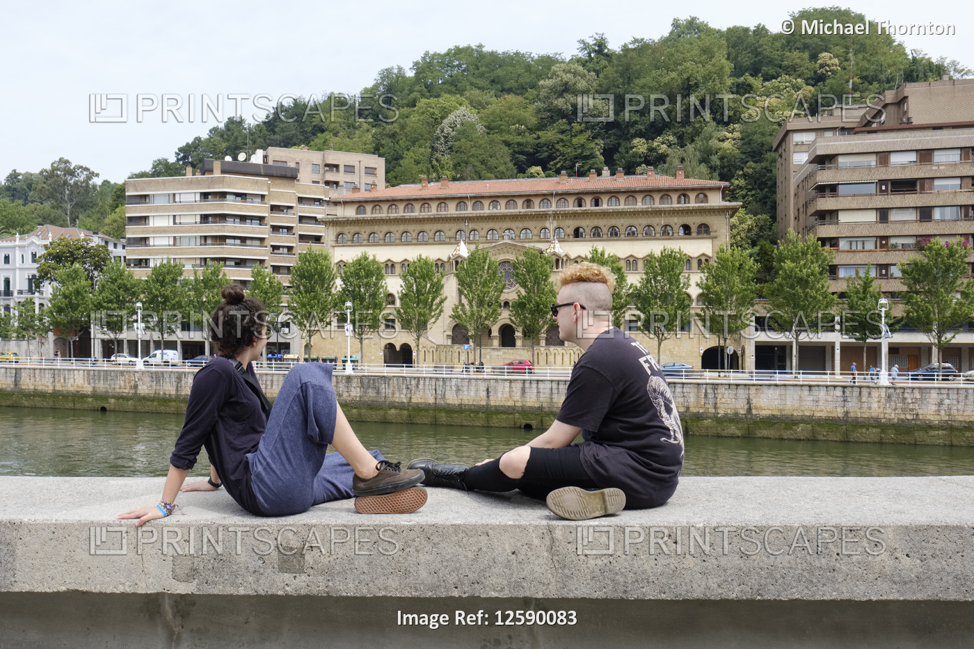 Young couple sitting on wall by the River Nervion, outside the Guggenheim ...