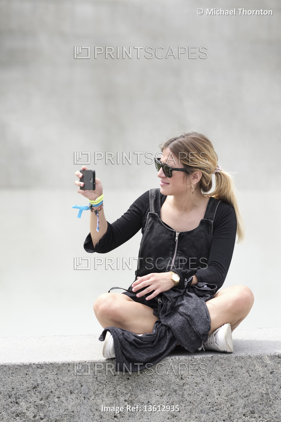 Young blonde lady with Ponytail, sitting on wall with mobile phone, taking a ...