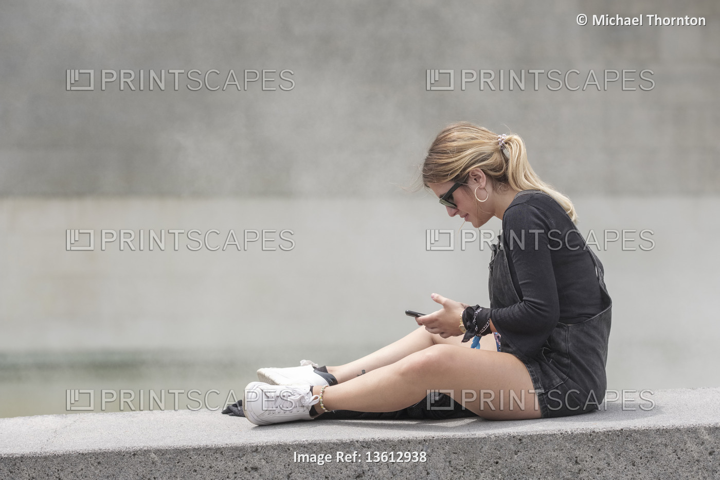 Young blonde lady with Ponytail, sitting on wall with mobile phone, Bilbao, ...